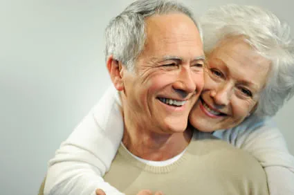 Ein älteres Ehepaar, welches lächelt und die Ehefrau den Ehemann von hinten umarmt