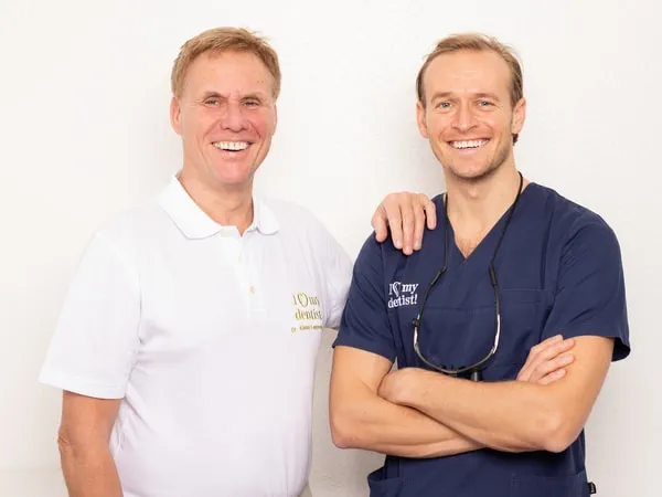 Die Zahnärzte Dr. Klaus und Alexander Langenbach stehen neben nebeneinander und lächeln